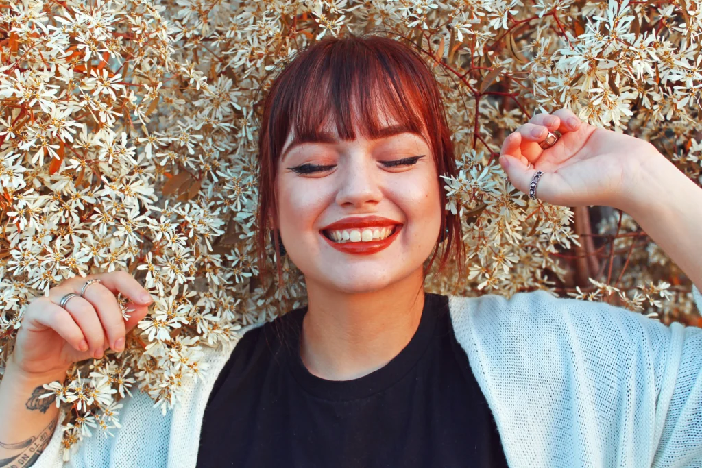 Portrait of a joyful woman with dark hair, wearing a black t-shirt and white cardigan, with her hands up near her head among delicate white blossoms.
