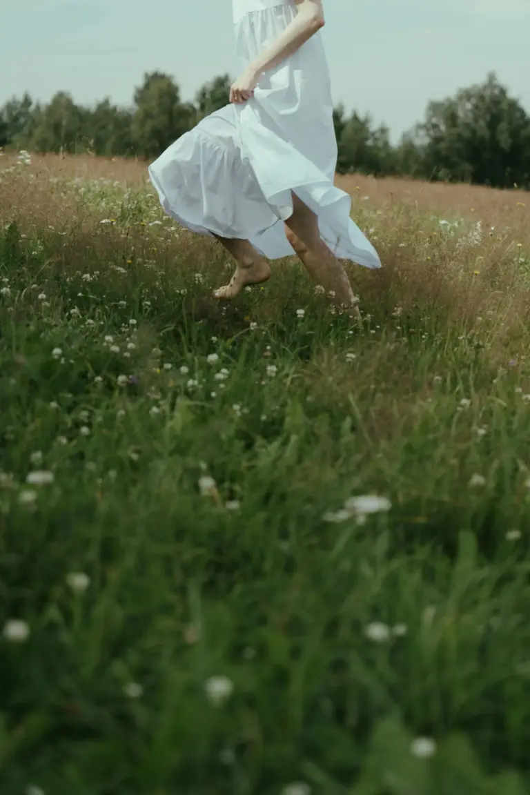 A barefoot woman in a flowing white dress running through a wildflower meadow, symbolizing natural beauty, freedom, and wellness.