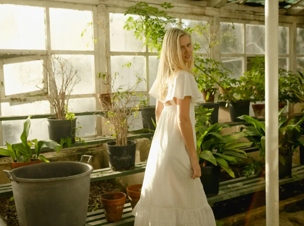 Woman in a white dress standing in a sunlit greenhouse surrounded by potted green plants