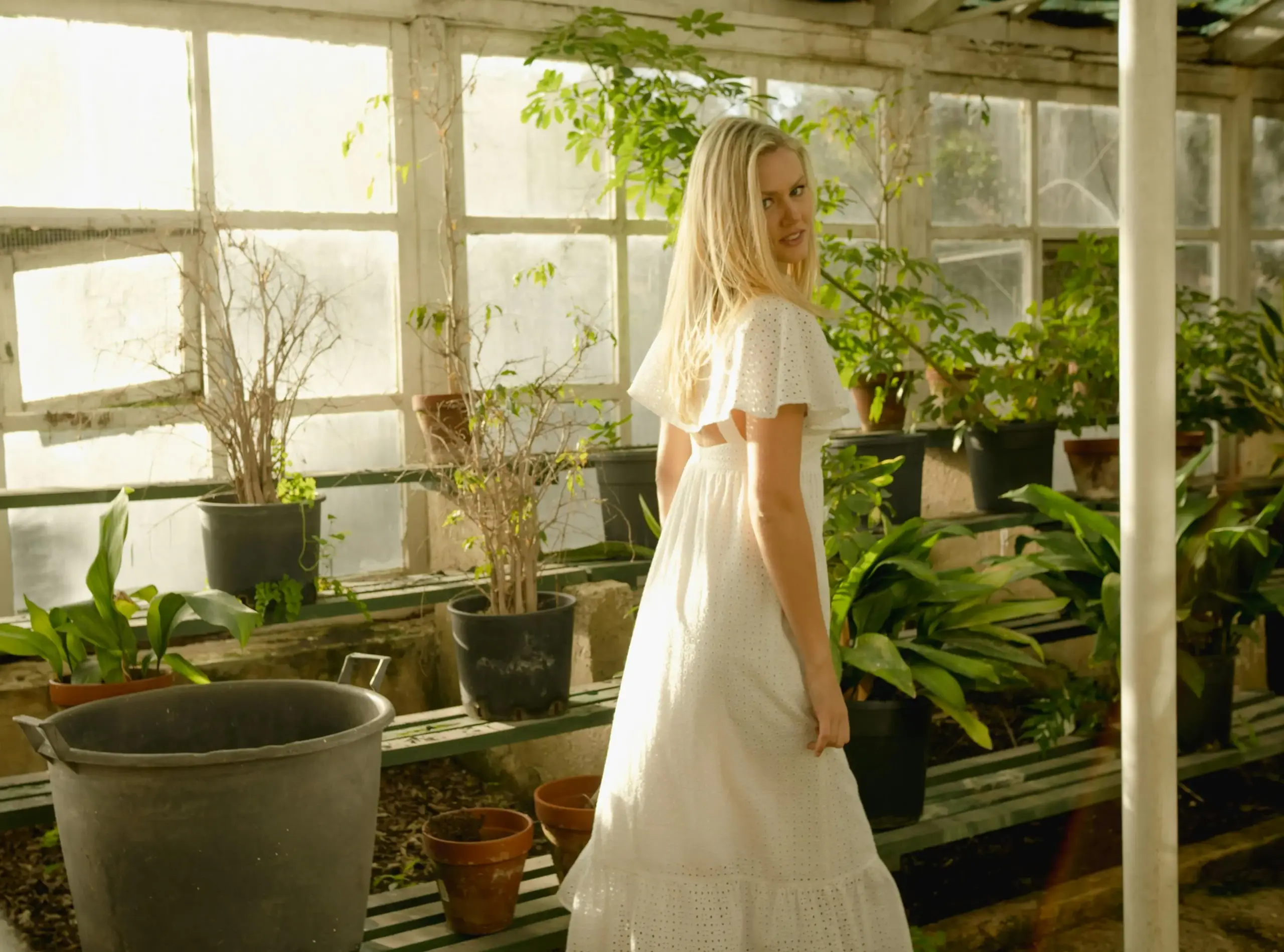 Woman in a white dress standing in a sunlit greenhouse surrounded by potted green plants
